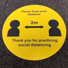 A yellow social distance floor marker circle with the words &rsquo; please keep your distance, thank you for practicing social distancing&rsquo; and a black and white outline of two stylized people and a double headed arrow between them which has 2m above it. 